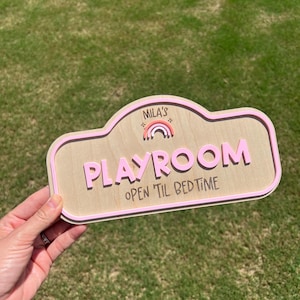 Puede incluir: Un letrero de madera con un borde rosa claro que presenta el texto "MILA'S" sobre un peque&ntilde;o gr&aacute;fico de arco&iacute;ris, la palabra "PLAYROOM" en letras rosas grandes y el texto "OPEN TIL BEDTIME" en negro, sobre un fondo de madera natural.