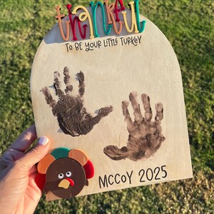 Puede incluir: Cartel de madera con la palabra "thankful" en letras coloridas. El cartel tiene dos huellas de manos y el texto "To be your little turkey" y "McCoy 2025". Una decoración de pavo está adjunta al cartel.