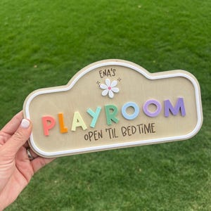 Puede incluir: Un letrero de madera clara con borde blanco que muestra el texto ENA'S PLAYROOM y OPEN 'TIL BEDTIME. La palabra PLAYROOM est&aacute; formada por letras en relieve de varios colores, incluyendo rojo, naranja, turquesa, azul y morado, con una flor blanca.
