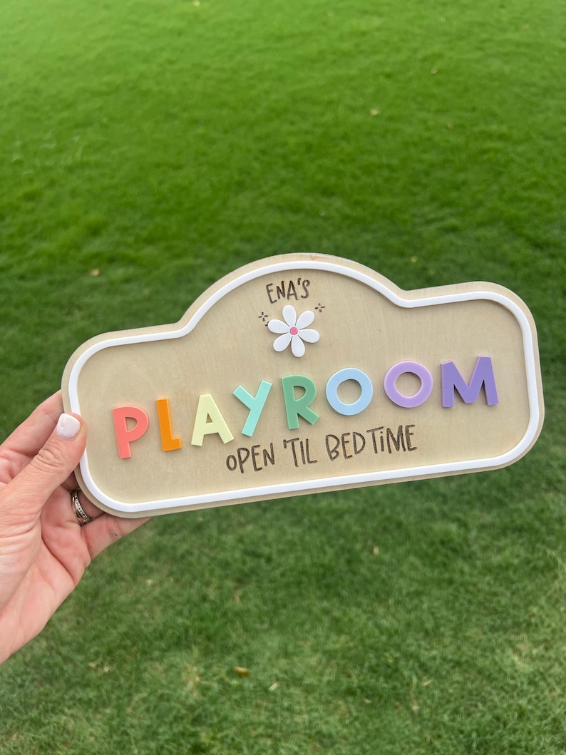 Puede incluir: Un letrero de madera clara con borde blanco que muestra el texto ENA'S PLAYROOM y OPEN 'TIL BEDTIME. La palabra PLAYROOM est&aacute; formada por letras en relieve de varios colores, incluyendo rojo, naranja, turquesa, azul y morado, con una flor blanca.