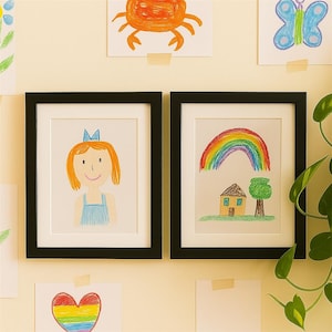 May include: Two black rectangular picture frames with white matting, each containing a child's crayon drawing. The left frame shows a person with orange hair and a blue dress. The right frame displays a colorful rainbow over a house and tree.