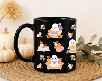 May include: Black ceramic mug decorated with whimsical ghost illustrations reading books, surrounded by autumn leaves, pumpkins, and candles. The mug has a large handle and is set on a woven mat, with pumpkins and flowers in the background.