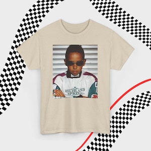 A sand t-shirt laid flat against a dynamic background of black-and-white checkered patterns and curved red lines. Centered on the shirt is a nostalgic photo print of a young Lewis Hamilton wearing sunglasses, arms crossed, and a vintage racing jacket with "Highland Spring" text. The brand name "VELORA" is displayed in the top-left corner in black, bold sans-serif font.