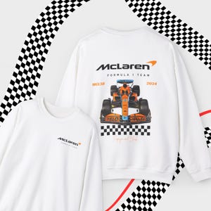 Puede incluir: Dos sudaderas blancas de manga larga con el logotipo del equipo McLaren de F&oacute;rmula 1. Una sudadera presenta un gr&aacute;fico de un coche de carreras McLaren con el texto "MCL38" y "2024".