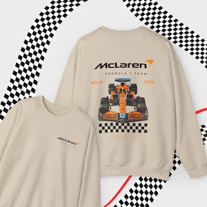 Puede incluir: Una sudadera beige con un logotipo de McLaren Formula 1 Team en la parte delantera y un logotipo m&aacute;s grande con un coche de carreras en la parte trasera. El logotipo de la parte trasera incluye el texto "McLaren Formula 1 Team", "MCL36" y "2024".