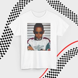 A white t-shirt laid flat against a dynamic background of black-and-white checkered patterns and curved red lines. Centered on the shirt is a nostalgic photo print of a young Lewis Hamilton wearing sunglasses, arms crossed, and a vintage racing jacket with "Highland Spring" text. The brand name "VELORA" is displayed in the top-left corner in black, bold sans-serif font.
