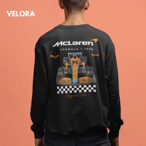 Puede incluir: Sudadera negra de manga larga con un logotipo de McLaren Formula 1 Team que presenta un coche de carreras naranja papaya y azul con el texto "McLaren Formula 1 Team MCL36 2024 Papaya and Black"