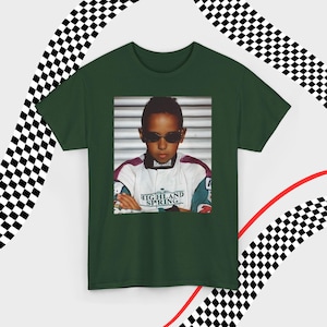 A green t-shirt laid flat against a dynamic background of black-and-white checkered patterns and curved red lines. Centered on the shirt is a nostalgic photo print of a young Lewis Hamilton wearing sunglasses, arms crossed, and a vintage racing jacket with "Highland Spring" text. The brand name "VELORA" is displayed in the top-left corner in black, bold sans-serif font.