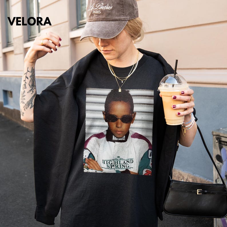 Woman wearing a black t-shirt with a printed childhood photo of Lewis Hamilton in racing gear and sunglasses, holding a drink, outdoors.