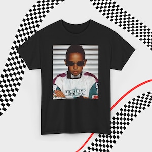 A black t-shirt laid flat against a dynamic background of black-and-white checkered patterns and curved red lines. Centered on the shirt is a nostalgic photo print of a young Lewis Hamilton wearing sunglasses, arms crossed, and a vintage racing jacket with "Highland Spring" text. The brand name "VELORA" is displayed in the top-left corner in black, bold sans-serif font.