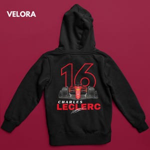 Puede incluir: Sudadera con capucha negra con un gr&aacute;fico de coche de carreras rojo y blanco en la parte posterior. El coche tiene el n&uacute;mero 16 y el texto "CHARLES LECLERC" con una firma.