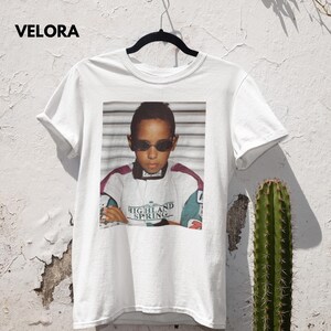A white t-shirt hanging on a black hanger in front of an old, textured white wall. The shirt features a nostalgic photo print of a young Lewis Hamilton, the future F1 champion, wearing sunglasses and a vintage racing jacket with "Highland Spring" text. To the immediate right of the shirt, a cactus plant is visible. The wall texture and the cactus add a rustic and authentic feel to the scene.