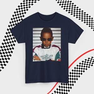 A navy t-shirt laid flat against a dynamic background of black-and-white checkered patterns and curved red lines. Centered on the shirt is a nostalgic photo print of a young Lewis Hamilton wearing sunglasses, arms crossed, and a vintage racing jacket with "Highland Spring" text. The brand name "VELORA" is displayed in the top-left corner in black, bold sans-serif font.