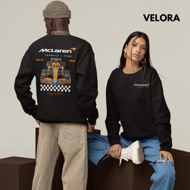 Puede incluir: Dos personas vistiendo sudaderas negras con el logotipo del equipo McLaren Formula 1 y el texto "McLaren Formula 1 Team 2023" y "McLaren Formula 1 Team". La persona de la izquierda lleva pantalones beige y la persona de la derecha lleva pantalones vaqueros azules.
