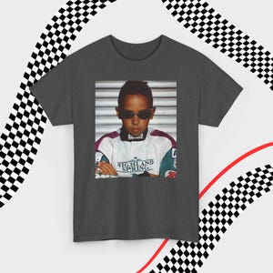 A gray t-shirt laid flat against a dynamic background of black-and-white checkered patterns and curved red lines. Centered on the shirt is a nostalgic photo print of a young Lewis Hamilton wearing sunglasses, arms crossed, and a vintage racing jacket with "Highland Spring" text. The brand name "VELORA" is displayed in the top-left corner in black, bold sans-serif font.