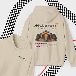 May include: A beige hoodie with a McLaren Formula 1 Team logo and a graphic of a race car. The hoodie also features a small Union Jack flag and the text "NORRIS".