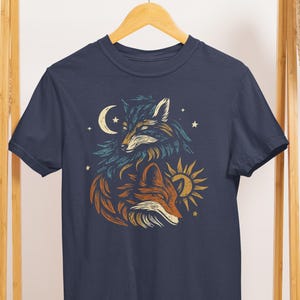 Op de afbeelding: Marineblauw t-shirt met een gestileerde illustratie van twee vossen, een met een maan en de andere met een zon. Het ontwerp gebruikt blauwe, oranje en gele tinten. Het t-shirt hangt aan een houten kledingrek.