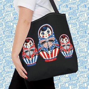 May include: Black tote bag featuring three Matryoshka dolls with patriotic red, white, and blue designs. The dolls have star and stripe patterns, with a bow detail. The bag has a shoulder strap and is held by a person.