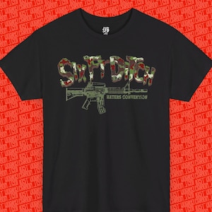Può includere: Una t-shirt nera a maniche corte con una stampa grafica al centro. Il design include il testo SIXFTDITCH in un motivo mimetico sopra l'illustrazione di un fucile e il testo HATERS CONVENTION in un carattere più piccolo verde oliva.