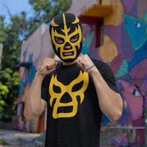 May include: Black t-shirt with a yellow luchador mask design. The person is wearing a matching black and yellow luchador mask. The mask and shirt feature a stylized design.