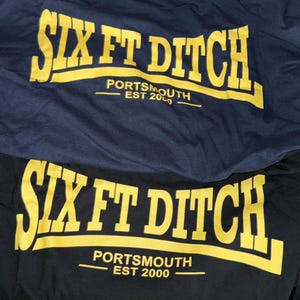 Puede incluir: Dos camisetas, una azul marino y otra negra, con el texto amarillo "SIX FT DITCH". Debajo, se lee "PORTSMOUTH EST 2000". Las camisetas están dobladas, mostrando el diseño.