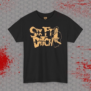 Peut inclure: Un t-shirt noir à manches courtes avec une impression graphique beige sur la poitrine. Le motif affiche le texte Six Ft Ditch dans une police dentelée à côté d'une illustration d'une silhouette tenant une tronçonneuse. L'étiquette intérieure indique SFD.