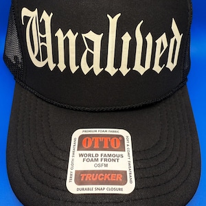 May include: Black trucker hat with the word "Unalived" in a gothic font. The hat features a mesh back and a curved brim. The front of the hat has a tag that reads "OTTO World Famous Foam Front OSFM Trucker".