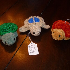 Puede incluir: Tres figuras de tortugas hechas a mano con ganchillo en verde, beige y naranja. Las tortugas tienen caparazones detallados y caras bonitas. Una tortuga tiene un diseño de caparazón azul. Una etiqueta dice "NAE'S COZY CRAFT HOUSE".