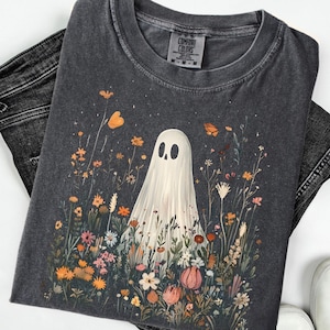 May include: Dark gray t-shirt featuring a whimsical ghost design amidst a colorful floral field. The ghost has a simple, friendly face. The shirt is paired with black jeans and white sneakers, creating a casual, everyday look.