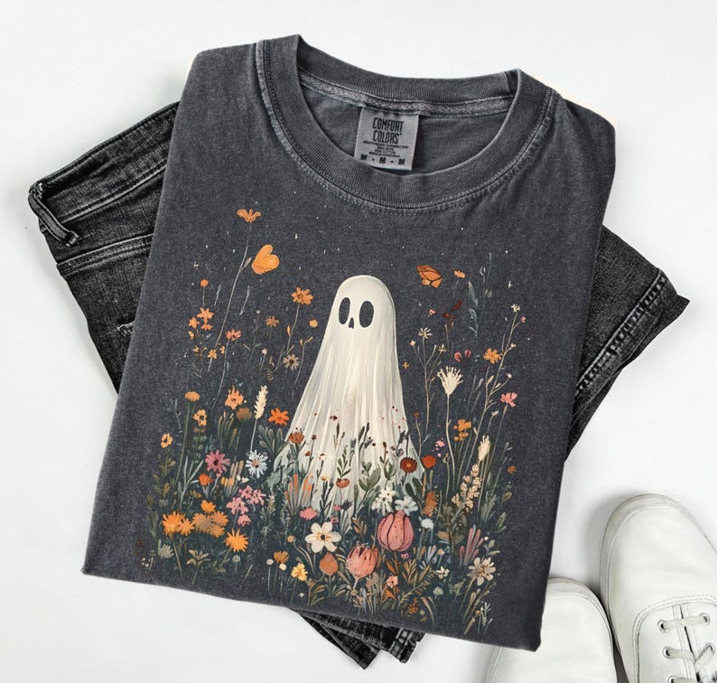 May include: Dark gray t-shirt featuring a whimsical ghost design amidst a colorful floral field. The ghost has a simple, friendly face. The shirt is paired with black jeans and white sneakers, creating a casual, everyday look.