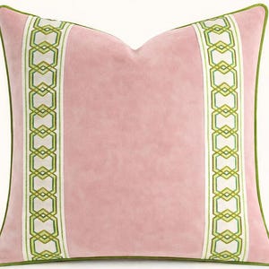 May include: A square decorative pillow in a soft pink hue. The pillow features two vertical bands with a geometric pattern in green and white. The edges are trimmed with a green border, creating a stylish accent for home decor.