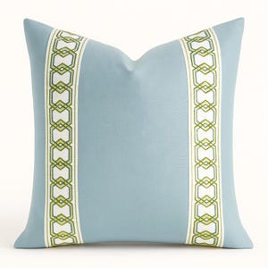 May include: A light blue decorative pillow with a geometric design. The pillow features two vertical bands of white fabric with a green geometric pattern. The pillow is a square shape and is made of a soft fabric.
