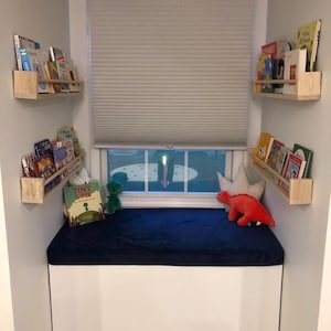 May include: A cozy reading nook with a white bench, a navy blue cushion, and a window with a closed blind. Wooden shelves hold children's books. Plush toys, including a green dinosaur and a red dinosaur, decorate the space.