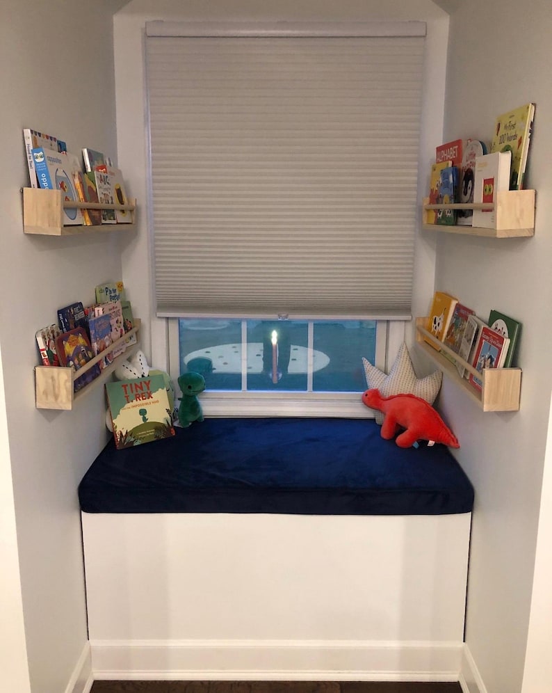 May include: A cozy reading nook with a white bench, a navy blue cushion, and a window with a closed blind. Wooden shelves hold children's books. Plush toys, including a green dinosaur and a red dinosaur, decorate the space.