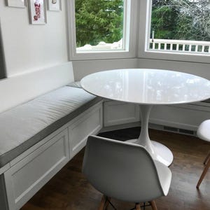May include: A white, round dining table with a single pedestal base. The table is paired with two white chairs and a built-in bench with a gray cushion. The room has a window with a view of trees.