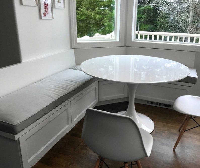 May include: A white, round dining table with a single pedestal base. The table is paired with two white chairs and a built-in bench with a gray cushion. The room has a window with a view of trees.