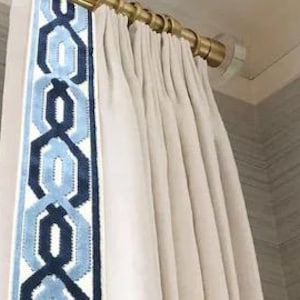May include: Cream-colored curtains with a blue and navy geometric border. The curtains are hanging from a gold-colored rod with clear, round finials. The fabric is pleated and hangs in soft folds.