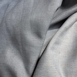 May include: Close-up of a light gray fabric with a woven texture. The fabric is draped, creating soft folds and shadows. The material appears to be a medium-weight textile, possibly linen or a similar natural fiber.