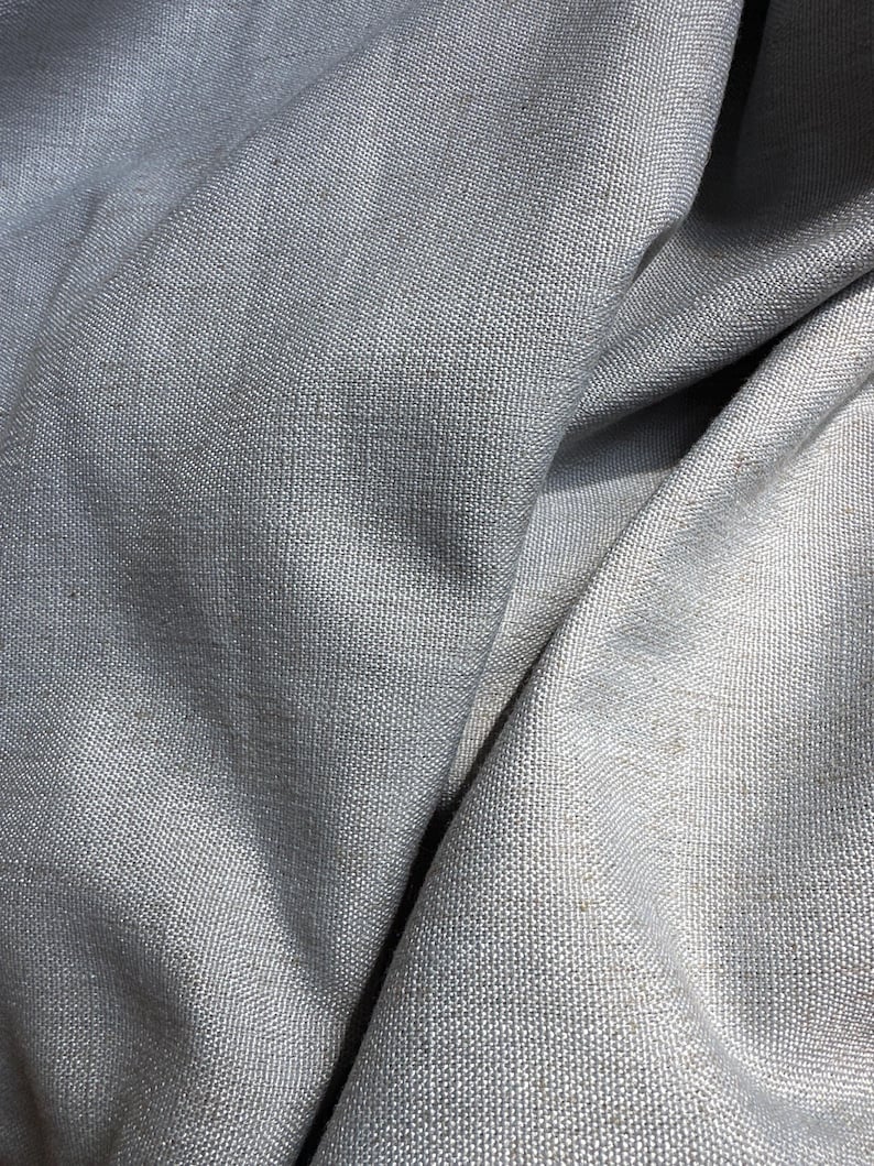May include: Close-up of a light gray fabric with a woven texture. The fabric is draped, creating soft folds and shadows. The material appears to be a medium-weight textile, possibly linen or a similar natural fiber.