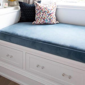 May include: A built-in window seat with a blue velvet cushion and white drawers. Two decorative pillows rest on the seat. The drawers have crystal knobs. The window seat is built into a white wall.