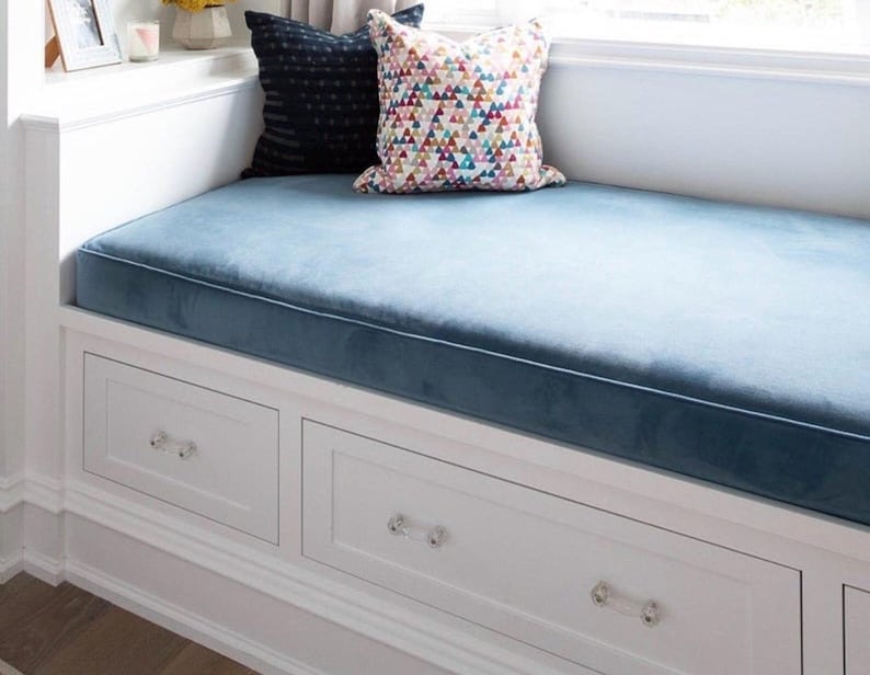May include: A built-in window seat with a blue velvet cushion and white drawers. Two decorative pillows rest on the seat. The drawers have crystal knobs. The window seat is built into a white wall.