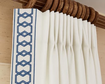 May include: Close-up of a white curtain with a blue geometric border. The curtain is pleated and hangs from a wooden rod with large, round wooden rings. The border features a repeating pattern of interlocking shapes.