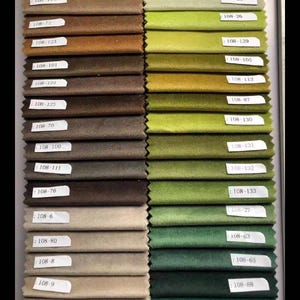 May include: A color swatch display featuring a variety of fabric samples in shades of green, brown, and beige. Each sample is labeled with a unique code, such as "108-24" and "108-122". The samples are arranged in neat rows, showcasing a range of textures and hues.