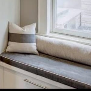 May include: A window seat with a gray cushion and a white and gray striped pillow. The cushion has a dark gray border. A long, cylindrical bolster pillow rests against the window frame.