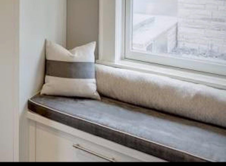 May include: A window seat with a gray cushion and a white and gray striped pillow. The cushion has a dark gray border. A long, cylindrical bolster pillow rests against the window frame.
