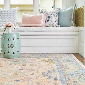 May include: A light blue ceramic garden stool with cut-out geometric designs sits near a window seat with decorative pillows. A floral patterned rug in peach, blue, and green tones covers the floor, creating a cozy interior design.