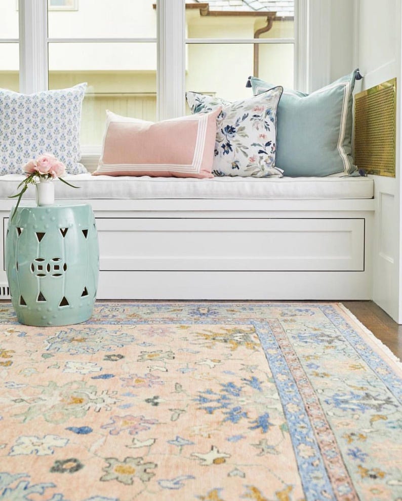 May include: A light blue ceramic garden stool with cut-out geometric designs sits near a window seat with decorative pillows. A floral patterned rug in peach, blue, and green tones covers the floor, creating a cozy interior design.
