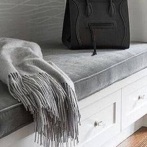 May include: A black leather handbag with a top handle and zipper detail sits on a gray cushion. A gray fringed throw blanket rests on the cushion next to the bag. The cushion is on a white bench with drawers.
