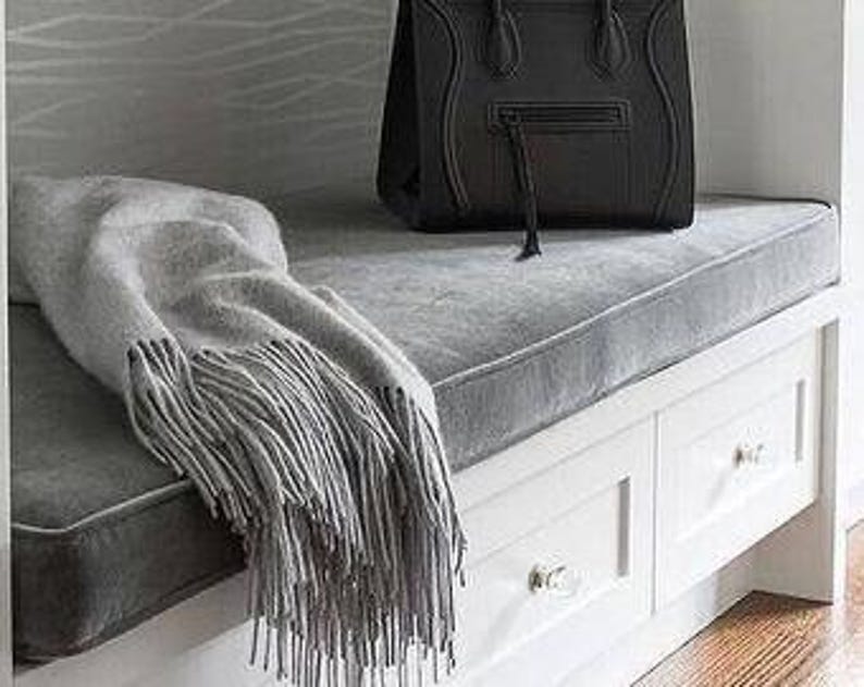 May include: A black leather handbag with a top handle and zipper detail sits on a gray cushion. A gray fringed throw blanket rests on the cushion next to the bag. The cushion is on a white bench with drawers.