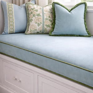 May include: A window seat with a light blue cushion and pillows. The pillows have decorative trim and patterns, including floral and geometric designs. The seat has a white storage base with two drawers.